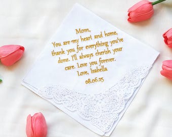 May include: White handkerchief with gold embroidered text: "Mom, You are my heart and home...Love, Isabella 08.06.25." Features a delicate lace trim. Pink tulips are scattered around the handkerchief, creating a soft, romantic scene.