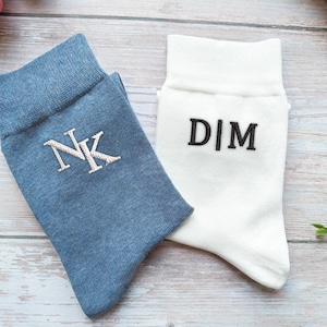 Può includere: Due paia di calzini, uno blu e uno bianco, con iniziali ricamate. Il calzino blu mostra "NK" in bianco, mentre il calzino bianco presenta "D|M" in nero. I calzini sono realizzati in materiale morbido.