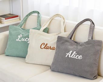 May include: Three corduroy tote bags in green, cream, and grey, each embroidered with a name: "Lucy", "Clara", and "Alice". The bags have matching handles and are displayed on a white sofa. The names are in contrasting colours.