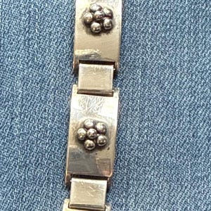 May include: Silver bracelet featuring rectangular links, each decorated with a cluster of small, round, raised floral designs. The bracelet is shown against a blue denim background.