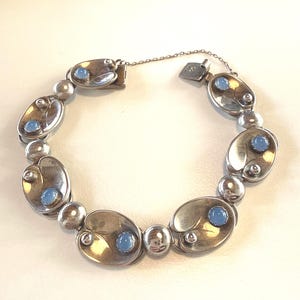 Peut inclure: Bracelet en argent composé de maillons ovales ornés de pierres cabochons bleu clair et de petits détails argentés. Le bracelet est doté d'un fermoir et d'une chaîne de sécurité. Un bijou classique.