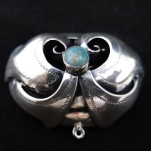 May include: A silver pendant with a central, round, pale blue stone. The pendant has a symmetrical design with curved, wing-like elements and decorative detailing. A small loop hangs from the bottom.