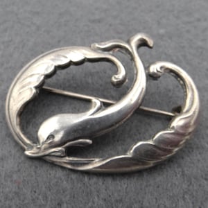 May include: A silver brooch depicting a dolphin leaping through stylised waves. The dolphin is the central element, with a curved body and detailed fins. The brooch has a pin closure on the back and is set against a grey background.