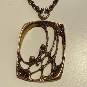 Puede incluir: Un collar con colgante vintage en tono dorado. El colgante tiene un marco rectangular abierto con un diseño abstracto que presenta formas orgánicas y pequeñas esferas. La cadena del collar es de color dorado a juego.