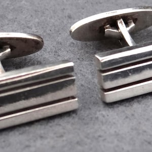 以下が含まれることがあります： 銀製のカフスボタンのペア。各カフスボタンは、3つの水平溝を備えた長方形のデザインが特徴です。接続ポストと背面は円形です。カフスボタンは灰色の表面に表示されています。