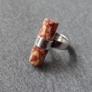 May include: A ring featuring a cylindrical stone bead with a marbled pattern of red, orange, and cream colours. The stone is held in place by a polished silver-toned metal band that wraps around the centre of the cylinder, creating a horizontal orientation.