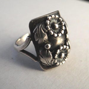 May include: A silver ring with a rectangular face adorned with floral and leaf motifs. Two detailed flowers are central to the design, complemented by leaves and smaller embellishments. The ring has a polished finish and a plain band.