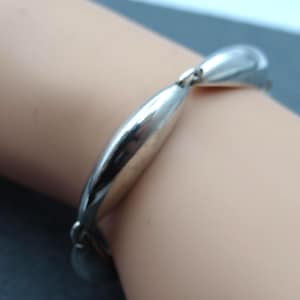 Puede incluir: Una pulsera plateada con una serie de eslabones ovalados y alargados. La pulsera es lisa y reflectante, con un cierre sencillo. La joya se muestra sobre un tono de piel claro.