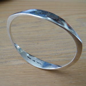 May include: A polished silver-toned bangle bracelet with a smooth, reflective surface and a slightly angular, geometric shape. The interior of the band features small, engraved text markings that are visible along the inner edge of the metal.