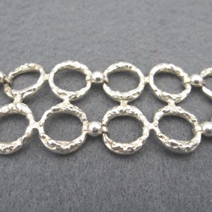 Puede incluir: Una pulsera plateada con un patrón repetitivo de círculos abiertos texturizados conectados por pequeñas cuentas redondas. La pulsera tiene un diseño único y orgánico, exhibida sobre un fondo gris.