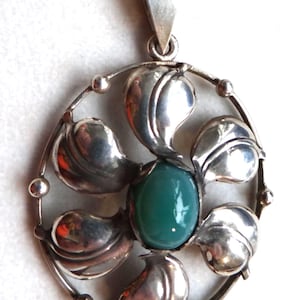 Może przedstawiać: Srebrny naszyjnik z wisiorkiem o kwiatowym wzorze. Wisiorek posiada centralny owalny zielony kamień otoczony srebrnymi elementami w kształcie liści. Naszyjnik ma delikatny łańcuszek.