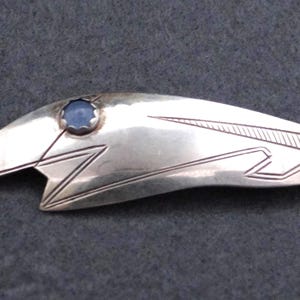Peut inclure: Broche en métal argenté au design stylisé. Elle présente une pierre bleue centrale et des motifs linéaires gravés. La forme générale est allongée et asymétrique, avec une esthétique épurée et moderne.