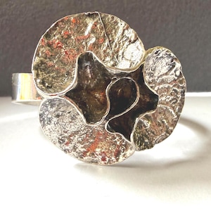 Puede incluir: Un anillo plateado con un diseño orgánico texturizado. El anillo presenta un elemento central en forma de estrella de color oscuro, rodeado por dos grandes piezas circulares texturizadas. La banda es de plata pulida simple.