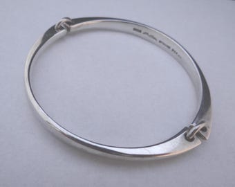 May include: A silver bracelet with a distinctive, curved shape. The bracelet has a smooth, polished surface and is joined by two decorative knots. The overall form is oval, and the metal has a bright, reflective appearance.