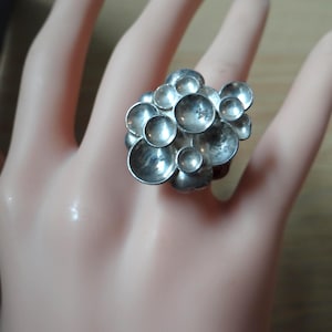 Peut inclure: Une bague argentée composée d'un groupe d'éléments circulaires concaves en forme de coupelles de tailles variées, disposés en une formation dense et organique. La surface métallique présente une finition polie avec des reflets.