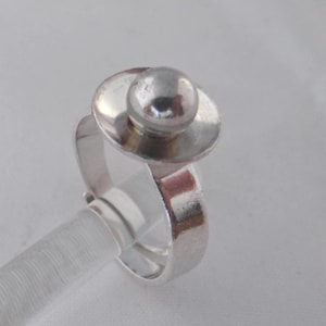 May include: A silver ring with a round, smooth, silver top. The ring is made of a thick band of silver.