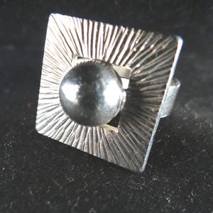 Puede incluir: Anillo plateado con una cara cuadrada con un diseño texturizado de rayos de sol. Una cuenta esférica oscura está centrada dentro del cuadrado. La banda del anillo es visible, lo que sugiere un tamaño de anillo estándar.