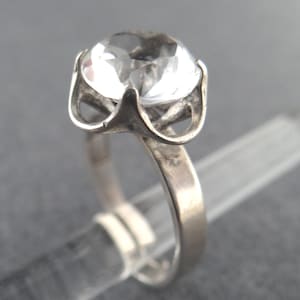 Könnte beinhalten: Ein Silberring mit einem großen, klaren, facettierten Edelstein. Der Edelstein ist rund und in einer Krappenfassung gefasst. Der Ring hat ein schlichtes, poliertes Silberband. Der Ring wird auf einem transparenten, zylindrischen Objekt vor grauem Hintergrund präsentiert.