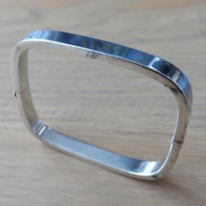 Peut inclure: Un bracelet jonc articulé de couleur argentée avec une forme rectangulaire aux coins arrondis. Le bracelet présente une surface métallique lisse et réfléchissante ainsi qu'une charnière visible sur le côté pour l'ouvrir et le fermer.