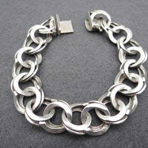 Könnte beinhalten: Ein silbernes Armband mit einer Reihe ineinandergreifender, abgerundeter Glieder. Das Armband ist geöffnet und zeigt den Verschlussmechanismus. Die Glieder sind dick und haben eine glatte, polierte Oberfläche. Das Armband liegt vor grauem Hintergrund.