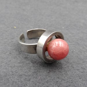 Puede incluir: Un anillo ajustable plateado con una gema rosa redonda y grande. El diseño del anillo incluye un elemento circular que acuna la gema, creando una estética moderna y minimalista. El anillo está sobre un fondo gris.