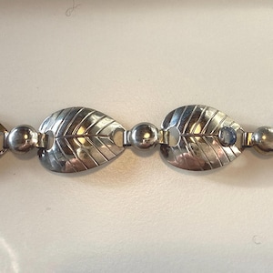 Peut inclure: Un bracelet argenté avec un motif de feuilles. Chaque maillon en forme de feuille présente des nervures détaillées et est relié par de petites perles rondes. De petites pierres bleues sont serties dans certaines des feuilles.