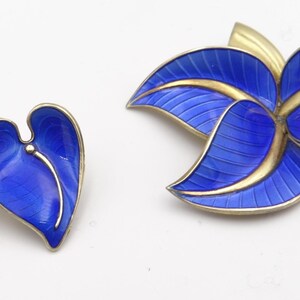 Peut inclure: Un ensemble de bijoux vintage comprenant une paire de boucles d'oreilles en forme de cœur en émail bleu et une broche fleur bleue et dorée. Les boucles d'oreilles et la broche ont une bordure en métal doré et une finition en émail bleu vif.