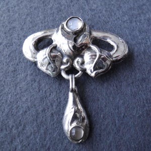Puede incluir: Broche plateado con diseño Art Nouveau. El broche presenta un motivo floral central y un colgante en forma de lágrima. Dos pequeñas piedras redondas y claras están engastadas en el broche y el colgante. El broche se muestra sobre un fondo gris.