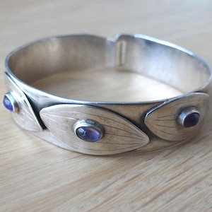 Könnte beinhalten: Silberfarbenes Armband mit Scharnier und drei blattförmigen Verzierungen, die jeweils mit einem kleinen, runden, lila Edelstein besetzt sind. Das Armband hat einen einfachen Verschluss und liegt auf einer hellen Holzoberfläche. Ein Schmuckstück.