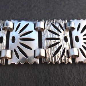 Pode incluir: Uma pulseira prateada com um padrão floral repetitivo. A pulseira apresenta padrões de raios de sol recortados e metal texturizado. As ligações são conectadas por pequenas barras cilíndricas. As bordas têm um design ondulado.