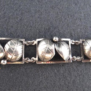 Peut inclure: Bracelet argenté avec un motif de feuilles répétitif. Chaque feuille est encadrée dans un rectangle, reliée par de petites chaînes. Les feuilles ont des nervures détaillées et de petites sphères ornent le design.