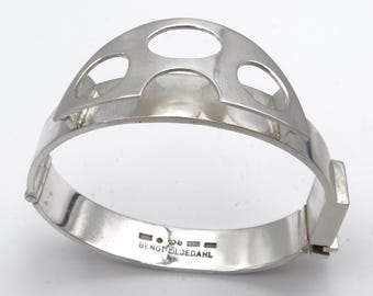 May include: A silver hinged bangle bracelet. The top section has a decorative cutout design with three oval openings. The bracelet is marked with "BENGT LILJEDAHL" and "925" on the inside. The bracelet is a piece of jewellery.