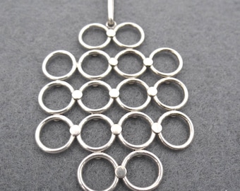Könnte beinhalten: Ein silberner Anhänger mit einem geometrischen Design aus miteinander verbundenen Kreisen. Der Anhänger hat eine Reihe von Doppelkreisen, die in einem Kaskadenmuster angeordnet und durch kleine, runde Akzente verbunden sind. Er hat eine Schlaufe zum Aufhängen.