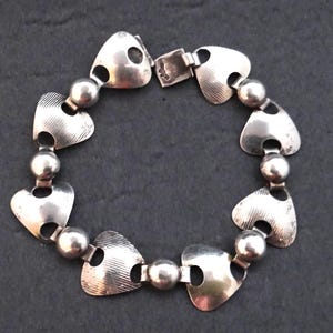 Peut inclure: Bracelet argenté composé de maillons en forme de cœur avec des détails texturés et de petites découpes circulaires. Les maillons sont reliés par des perles sphériques et un fermoir. Un bijou.