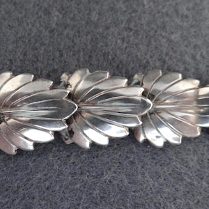 Peut inclure: Un bracelet argenté avec un motif de feuilles répétées. Chaque feuille présente des nervures détaillées et une finition polie. Le bracelet est présenté sur un fond gris, soulignant son éclat métallique.