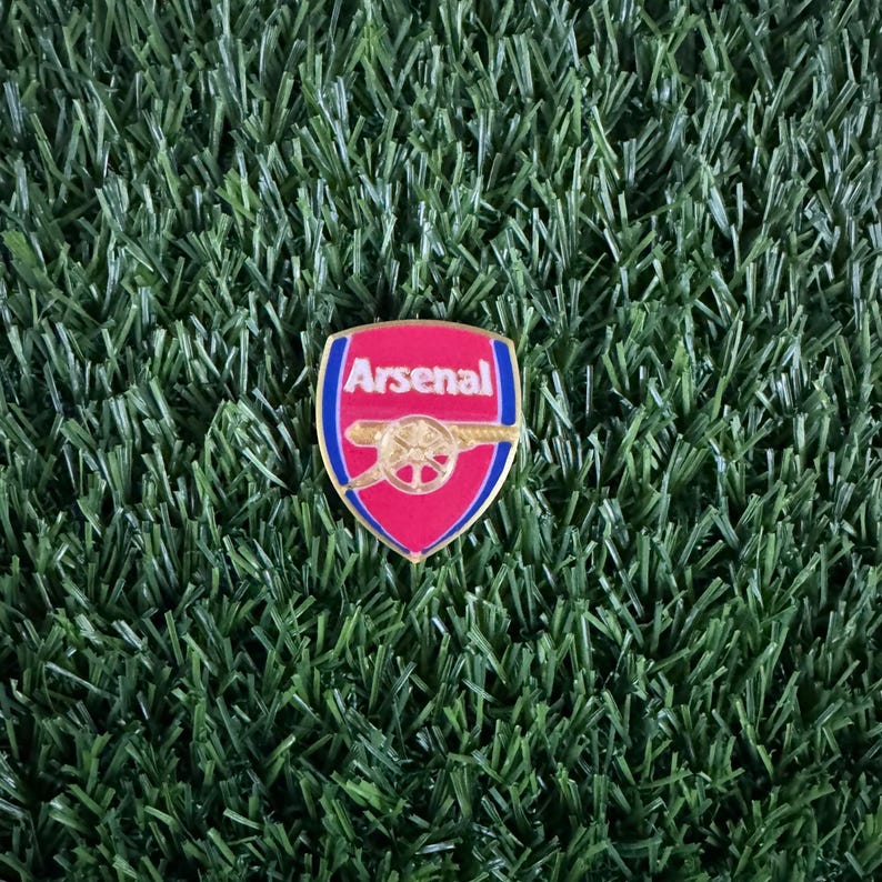 May include: A red and gold Arsenal football club pin with the team's logo and name.