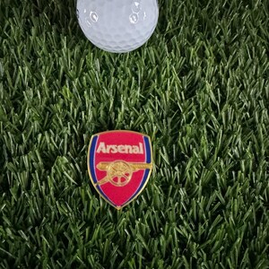 May include: A red, white, and blue Arsenal football club pin with a cannon graphic. The pin is on a green artificial grass surface.
