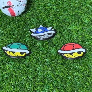 Op de afbeelding: Drie kleurrijke plastic golfbalmarkers in de vorm van schildpaddenpantsers. De markers zijn rood, groen en blauw. De rode en groene markers hebben een witte omtrek en een gele onderkant. De blauwe marker heeft een grijze onderkant en witte stekels.