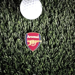 May include: A gold and red Arsenal football club pin with the team's name and crest. The pin is on a green artificial grass surface.