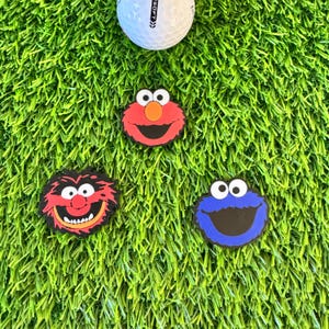 May include: Three golf ball markers featuring Sesame Street characters: Big Bird, Elmo, and Cookie Monster. The markers are red, blue, and orange.