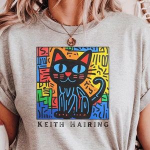 May include: Gray t-shirt with a colorful graphic of a black cat and the text "Keith Haring".