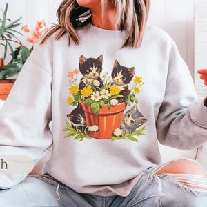 May include: A white sweatshirt with a graphic of three kittens in a flower pot. The kittens are black and white with green, yellow, and white flowers around them. The text "Ash" is in the bottom left corner of the image.