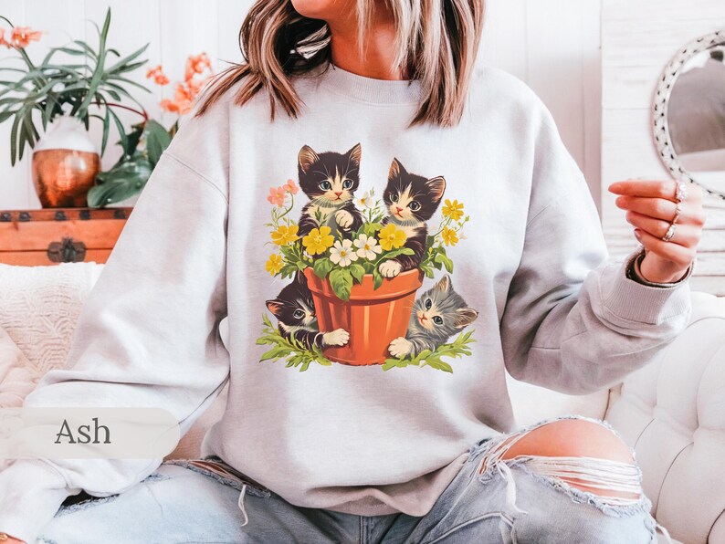 May include: A white sweatshirt with a graphic of three kittens in a flower pot. The kittens are black and white with green, yellow, and white flowers around them. The text "Ash" is in the bottom left corner of the image.