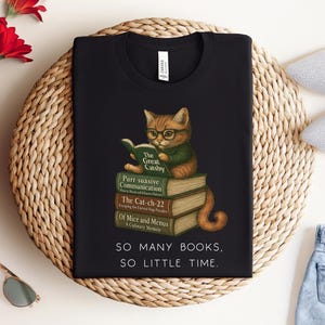 K&ouml;nnte beinhalten: Schwarzes T-Shirt mit einem Grafikdruck eines Katers, der einen Stapel B&uuml;cher liest. Der Text auf dem Shirt lautet "SO MANY BOOKS, SO LITTLE TIME."