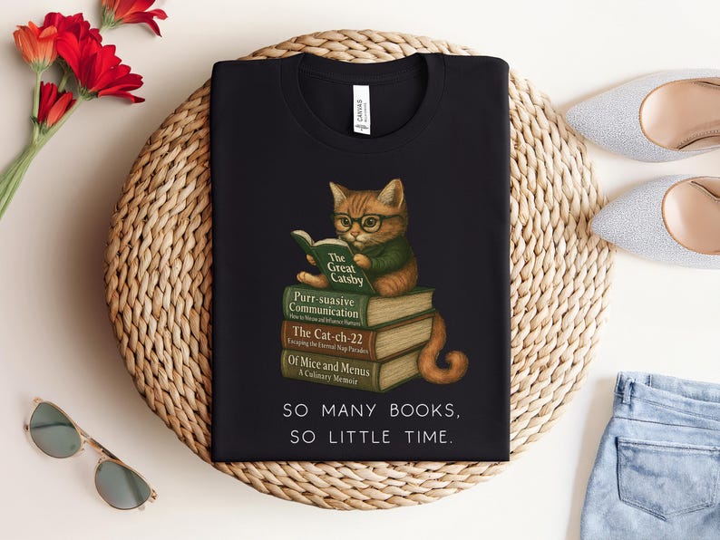 K&ouml;nnte beinhalten: Schwarzes T-Shirt mit einem Grafikdruck eines Katers, der einen Stapel B&uuml;cher liest. Der Text auf dem Shirt lautet "SO MANY BOOKS, SO LITTLE TIME."