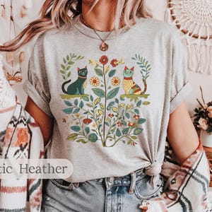 May include: A heather grey t-shirt with a floral design featuring two cats. The cats are stylized and have a folk art aesthetic. The design includes flowers, leaves, and stems.