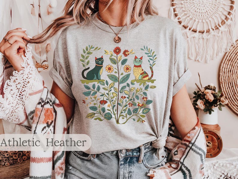 May include: A heather grey t-shirt with a floral design featuring two cats. The cats are stylized and have a folk art aesthetic. The design includes flowers, leaves, and stems.