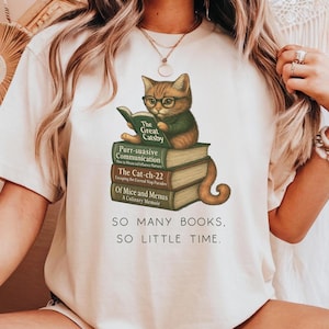 May include: A white t-shirt with a graphic of a cat wearing glasses and reading a book titled "The Great Gatsby". The cat is sitting on a stack of books with titles like "Purr-suasive Communication", "The Cat-ch-22", and "Of Mice and Menus". The text "SO MANY BOOKS, SO LITTLE TIME." is printed below the graphic.