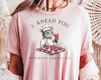 May include: Pink t-shirt with a graphic of a cat wearing a chef's hat and holding a heart-shaped cookie. The text "I knead you" is above the cat and "Whiskers Bakery Co." is below the cat.
