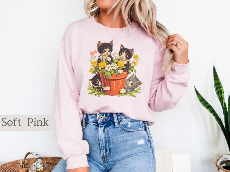 May include: A soft pink sweatshirt with a graphic of three kittens in a flower pot. The kittens are black and white with yellow flowers around them.